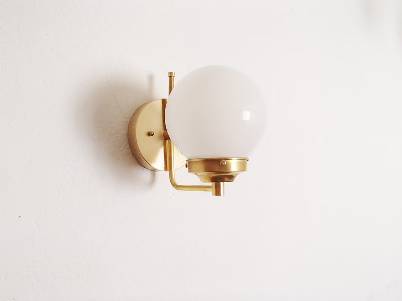 Peut inclure: Une applique murale dor&eacute;e avec un globe lumineux blanc. L'applique a une base ronde et un bras incurv&eacute;.