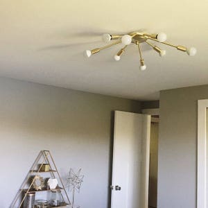 May include: A gold-toned ceiling light fixture with multiple arms extending outwards. Each arm has a white light bulb at the end. The light fixture is mounted on a white ceiling.