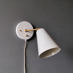 Peut inclure: Applique murale blanche avec un abat-jour conique et un bras en laiton. L'applique est équipée d'un cordon blanc.