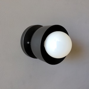 A modern and minimalist style wall sconce with half spherical shaped shade, flat wall base, dark oil rubbed bronze finish, white globe bulb. Gray background.