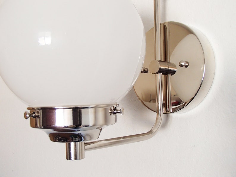 Peut inclure: Une applique murale chrom&eacute;e avec un abat-jour en verre blanc sph&eacute;rique. L'applique a un bras incurv&eacute; et une base ronde.