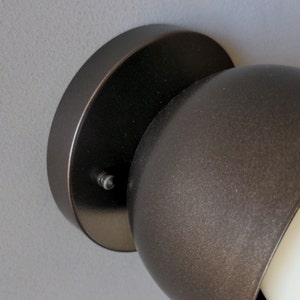 A modern and minimalist style wall sconce with half spherical shaped shade, flat wall base, dark oil rubbed bronze finish, white globe bulb. Gray background. Up close detailed photo.