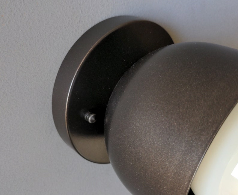 A modern and minimalist style wall sconce with half spherical shaped shade, flat wall base, dark oil rubbed bronze finish, white globe bulb. Gray background. Up close detailed photo.