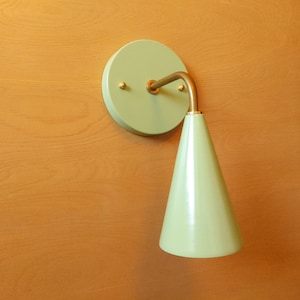 Lighting sconce, pastel mint green wall base and narrow tapered cone shade, with gold unfinished brass 90 degree curved stem and mounting hardware. Wood grain background.