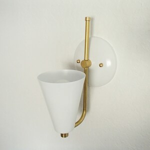 Puede incluir: Una l&aacute;mpara de pared blanca con un brazo de metal dorado y una pantalla c&oacute;nica blanca. La l&aacute;mpara tiene una placa trasera circular blanca con dos tornillos dorados.