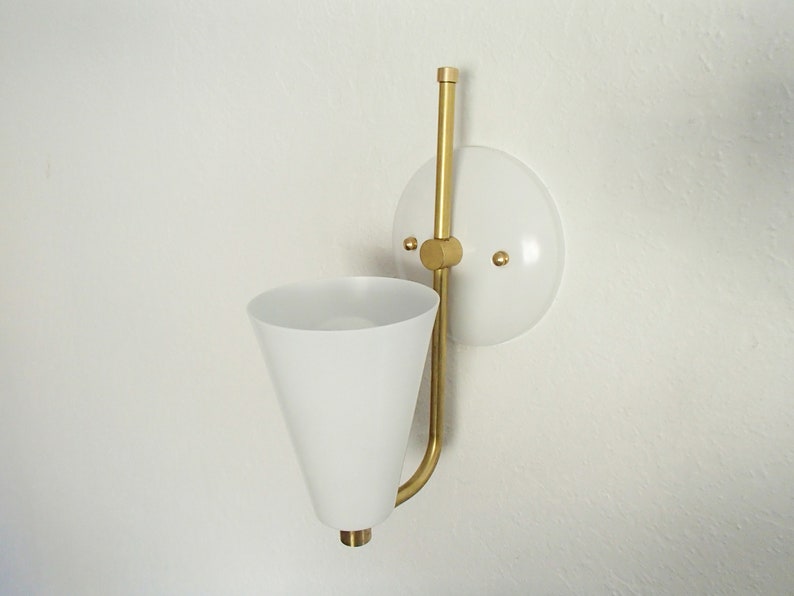 Puede incluir: Una l&aacute;mpara de pared blanca con un brazo de metal dorado y una pantalla c&oacute;nica blanca. La l&aacute;mpara tiene una placa trasera circular blanca con dos tornillos dorados.
