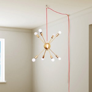 May include: A gold-toned, multi-arm chandelier with white light bulbs hangs from a red cord against a white ceiling. The chandelier is suspended in a room with a window and a doorway.
