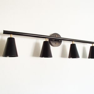 May include: Black metal light fixture with four black cone-shaped shades. The fixture is mounted to a wall and has a long, straight bar with the shades extending out from it.