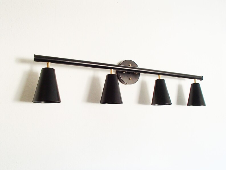 May include: Black metal light fixture with four black cone-shaped shades. The fixture is mounted to a wall and has a long, straight bar with the shades extending out from it.