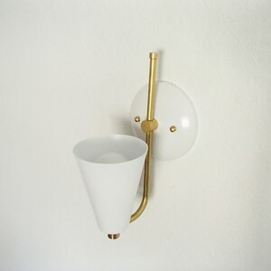 Puede incluir: Una l&aacute;mpara de pared blanca con un brazo de metal dorado y una pantalla c&oacute;nica blanca. La l&aacute;mpara est&aacute; montada en una placa circular blanca.