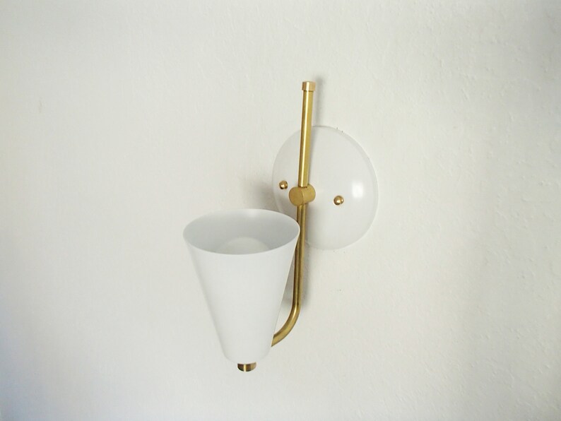Puede incluir: Una l&aacute;mpara de pared blanca con un brazo de metal dorado y una pantalla c&oacute;nica blanca. La l&aacute;mpara est&aacute; montada en una placa circular blanca.