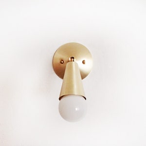 Satin brass finished wall lighting sconce. Round wall base, stem with swivel and small conical shade angled downwards. White globe bulb fitted. White background.