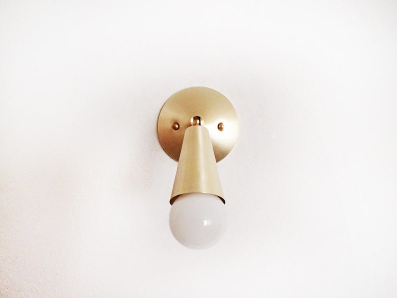 Satin brass finished wall lighting sconce. Round wall base, stem with swivel and small conical shade angled downwards. White globe bulb fitted. White background.