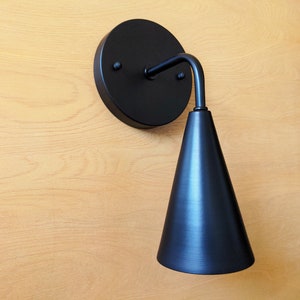 Lighting sconce, wall base and narrow tapered cone shade, with  90 degree curved stem and mounting hardware all black finish. Wood grain background.