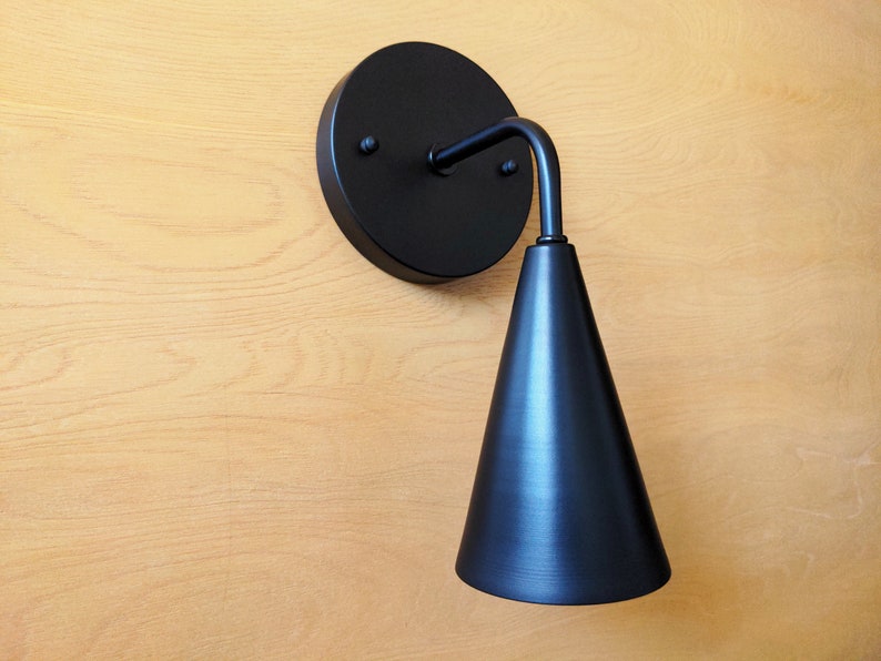 Lighting sconce, wall base and narrow tapered cone shade, with  90 degree curved stem and mounting hardware all black finish. Wood grain background.