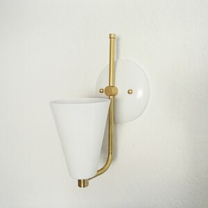 Puede incluir: Una l&aacute;mpara de pared blanca con una pantalla blanca en forma de cono y un brazo de metal dorado. La l&aacute;mpara est&aacute; montada en una base circular blanca.