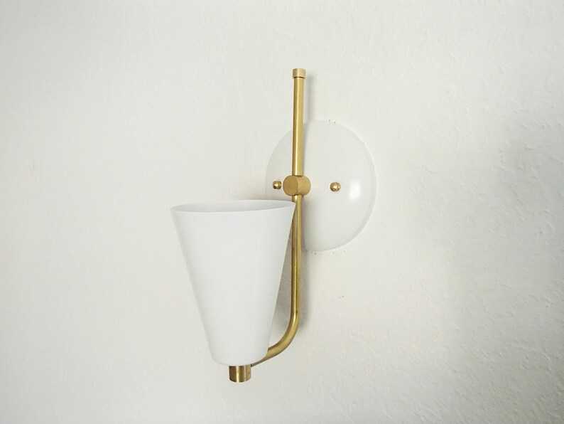 Puede incluir: Una l&aacute;mpara de pared blanca con una pantalla blanca en forma de cono y un brazo de metal dorado. La l&aacute;mpara est&aacute; montada en una base circular blanca.
