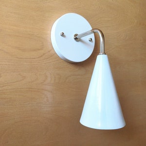 Lighting sconce, white wall base and narrow tapered cone shade, with bright polished nickel 90 degree curved stem and mounting hardware. Wood grain background.