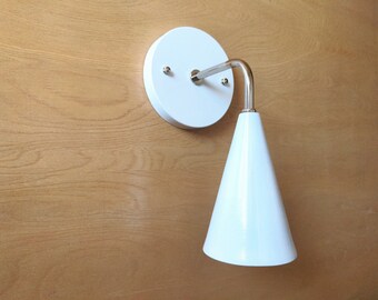 May include: White wall-mounted light fixture with a cone-shaped shade. The fixture has a white base and a silver arm.