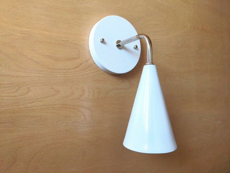 Lighting sconce, white wall base and narrow tapered cone shade, with bright polished nickel 90 degree curved stem and mounting hardware. Wood grain background.