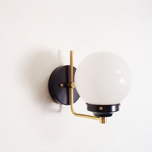 Peut inclure: Une applique murale noire et laiton avec un abat-jour globe blanc. L'applique a un design simple et moderne.