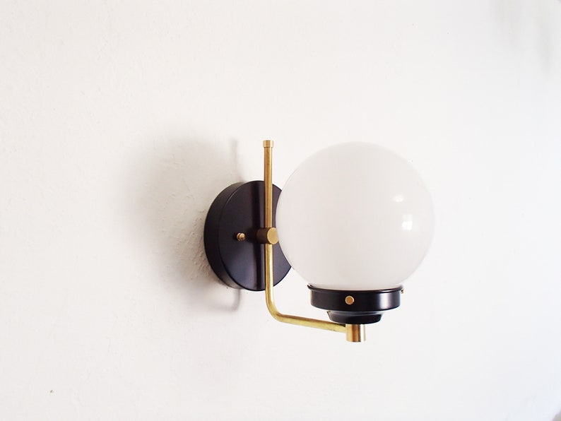 Peut inclure: Une applique murale noire et laiton avec un abat-jour globe blanc. L'applique a un design simple et moderne.