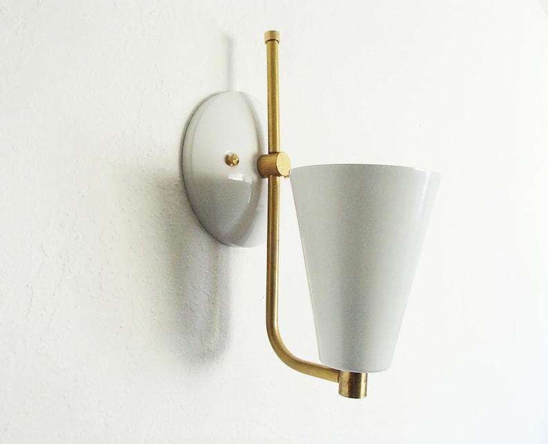 Puede incluir: Una l&aacute;mpara de pared blanca con un brazo de lat&oacute;n y una pantalla c&oacute;nica blanca.