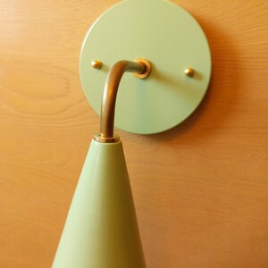 Lighting sconce, pastel mint green wall base and narrow tapered cone shade, with gold unfinished brass 90 degree curved stem and mounting hardware. Wood grain background.