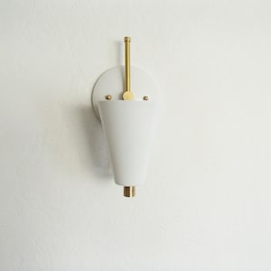 Puede incluir: Aplique de pared blanca en forma de cono con un v&aacute;stago de lat&oacute;n y una base redonda de lat&oacute;n. La aplique est&aacute; montada en una pared blanca.