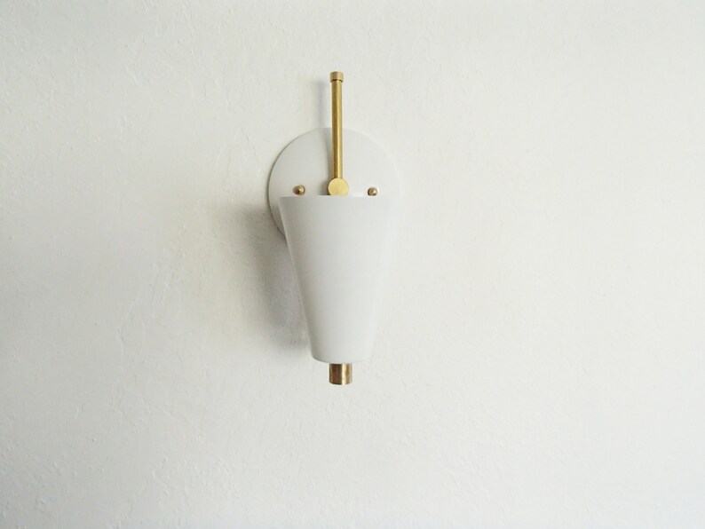 Puede incluir: Aplique de pared blanca en forma de cono con un v&aacute;stago de lat&oacute;n y una base redonda de lat&oacute;n. La aplique est&aacute; montada en una pared blanca.