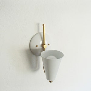 Puede incluir: Aplique de pared blanca con una pantalla en forma de cono y un brazo de lat&oacute;n. La aplique est&aacute; montada en una pared blanca.