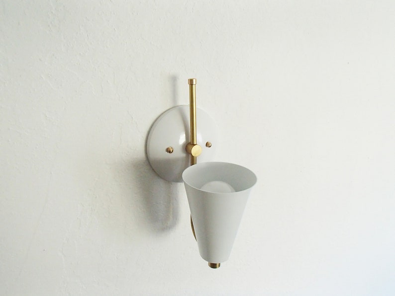 Puede incluir: Aplique de pared blanca con una pantalla en forma de cono y un brazo de lat&oacute;n. La aplique est&aacute; montada en una pared blanca.