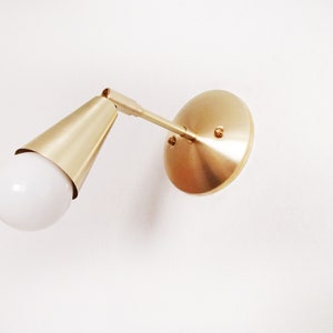 Satin brass finished wall lighting sconce. Round wall base, stem with swivel and small conical shade angled downwards. White globe bulb fitted. White background. View looking upwards.