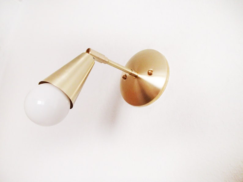 Satin brass finished wall lighting sconce. Round wall base, stem with swivel and small conical shade angled downwards. White globe bulb fitted. White background. View looking upwards.