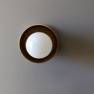 A modern and minimalist style wall sconce with half spherical shaped shade, flat wall base, bright metallic gold finish, white globe bulb. Gray background.