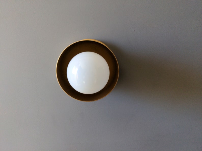 A modern and minimalist style wall sconce with half spherical shaped shade, flat wall base, bright metallic gold finish, white globe bulb. Gray background.