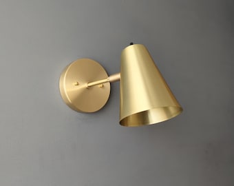 May include: A gold-toned wall sconce with a cone-shaped shade. The sconce has a round base and a single light bulb.
