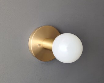 May include: A gold wall sconce with a white globe light bulb. The sconce has a round base and a short arm that extends out to the bulb.