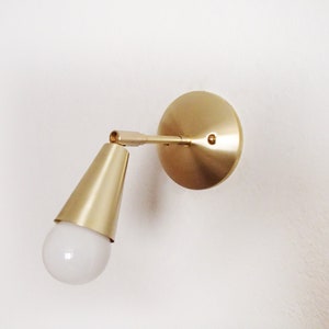 Satin brass finished wall lighting sconce. Round wall base, stem with swivel and small conical shade angled downwards. White globe bulb fitted. White background.