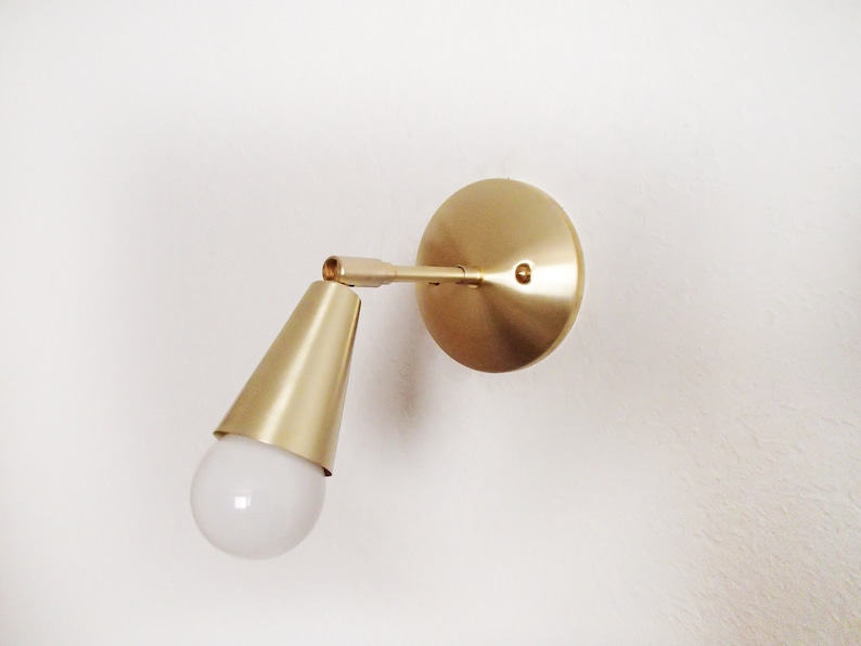 Satin brass finished wall lighting sconce. Round wall base, stem with swivel and small conical shade angled downwards. White globe bulb fitted. White background.