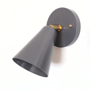 Può includere: Un'applique da parete in metallo grigio con un paralume a forma di cono e un accento in ottone. L'applique è montata su una parete bianca.