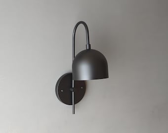 May include: A black metal wall sconce with a curved arm and a dome-shaped shade. The sconce is mounted on a white wall.