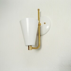 Puede incluir: Una pantalla de l&aacute;mpara blanca en forma de cono con un brazo y una base de metal dorado. La l&aacute;mpara est&aacute; montada en una pared blanca.
