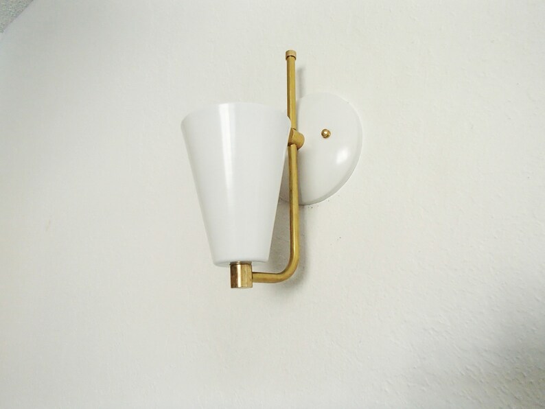 Puede incluir: Una pantalla de l&aacute;mpara blanca en forma de cono con un brazo y una base de metal dorado. La l&aacute;mpara est&aacute; montada en una pared blanca.