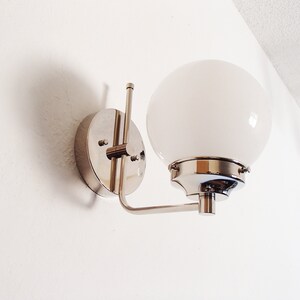 Peut inclure: Une applique murale chrom&eacute;e avec un luminaire blanc en forme de globe. L'applique a un design simple et moderne.