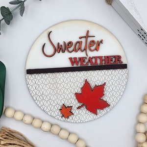 May include: A round wooden sign with the text "Sweater Weather" in orange and red lettering. The sign has a white background with a black and white knit pattern and two red and orange maple leaves.