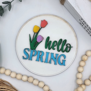 May include: A white round wooden sign with the words "hello spring" in green and blue lettering. The sign is decorated with three colourful tulips.