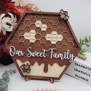 May include: A wooden honeycomb-shaped sign with the words "Our Sweet Family" and the names Mike, Jessica, Michael, Haily, Ethan, Aeris, Logan, Alexander, and Celes written in the honeycomb cells. The sign has a bee cutout on the top and bottom and is standing on a wooden base.