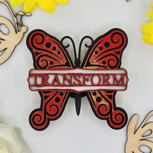 May include: A wooden butterfly with red and gold paint and black accents. The butterfly has the word "TRANSFORM" written on a white banner.