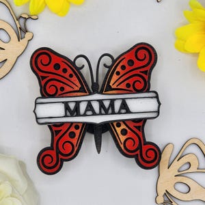 May include: A wooden butterfly with red and orange wings and black detailing. The butterfly has a white banner across its wings with the word "MAMA" written in black.